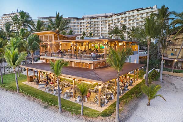 A beachside, two-story restaurant with outdoor seating is surrounded by palm trees and white sand at sunset, with a large Mexico hotel in the background. Diners enjoy the open-air atmosphere, perfect for relaxed meetings or conference events.