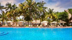 En uno de los complejos turísticos más exclusivos de México, una gran piscina al aire libre de agua cristalina está rodeada de tumbonas y sombrillas, con altas palmeras y cabañas al fondo: un entorno ideal para reuniones o conferencias bajo un cielo despejado.