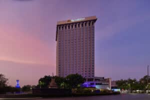 Un alto edificio de hotel con el letrero de Fiesta Americana en la parte superior, con un colorido cielo crepuscular de fondo, rodeado de árboles y un pequeño monumento, perfecto para hoteles en México que ofrecen eventos y reuniones, con una calle en primer plano.