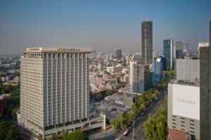 Vista aérea de un paisaje urbano con el prominente edificio Posada Reforma en primer plano, rodeado de otros hoteles mexicanos, espacios para eventos y conferencias, frondosos árboles verdes y un cielo azul despejado al fondo.