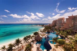 Un complejo turístico tropical en México con edificios altos, una piscina serpenteante rodeada de palmeras y una playa de arena blanca con vistas a aguas oceánicas turquesas: perfecto para conferencias o reuniones bajo cielos azules con nubes dispersas.