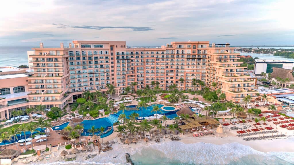 A large beachfront resort in Mexico with multiple stories, balconies, lush palm trees, several swimming pools, loungers, and umbrellas. Perfect for meetings and conference events, the hotel overlooks a sandy beach with waves and tropical landscaping.