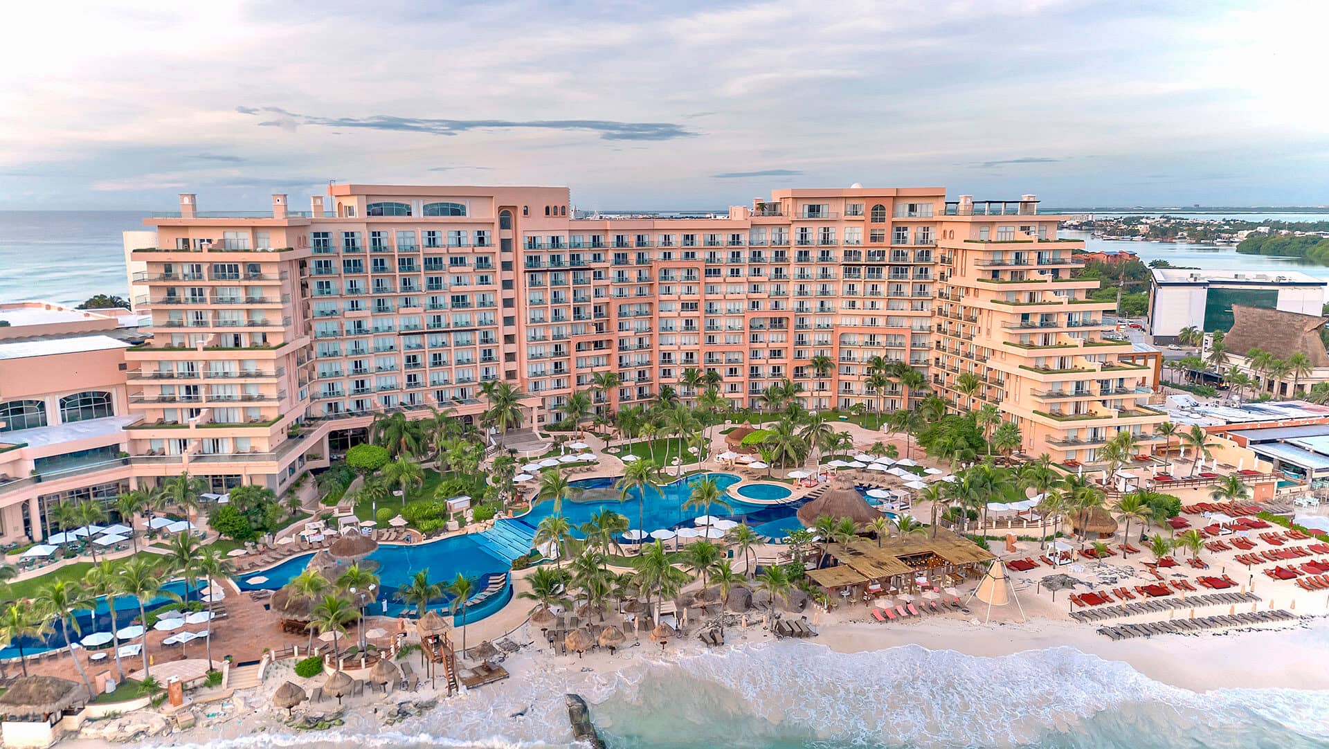 Un gran resort frente al mar en México, con varios pisos, balcones, exuberantes palmeras, varias piscinas, tumbonas y sombrillas. Ideal para reuniones y eventos, el hotel tiene vistas a una playa de arena con olas y jardines tropicales.