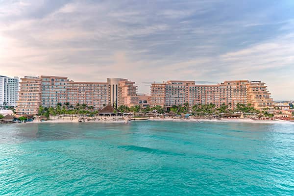 Large beachfront resort in Mexico with multiple buildings and balconies, ideal for conference events and meetings, surrounded by palm trees and facing turquoise ocean water under a partly cloudy sky.