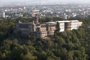 Un gran castillo histórico se alza sobre una colina boscosa con vistas a un extenso paisaje urbano a la luz del día, rodeado de densos árboles y edificios urbanos: un telón de fondo ideal para eventos de conferencias o reuniones en un complejo turístico.