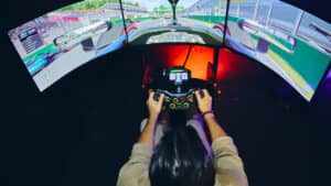 Una persona se sienta en un simulador de carreras con tres monitores curvos que muestran una pista de Fórmula 1, sosteniendo un volante; una experiencia que se suele encontrar en eventos de conferencias en los mejores hoteles y complejos turísticos de México, sumergiendo a los huéspedes en juegos de conducción realistas.