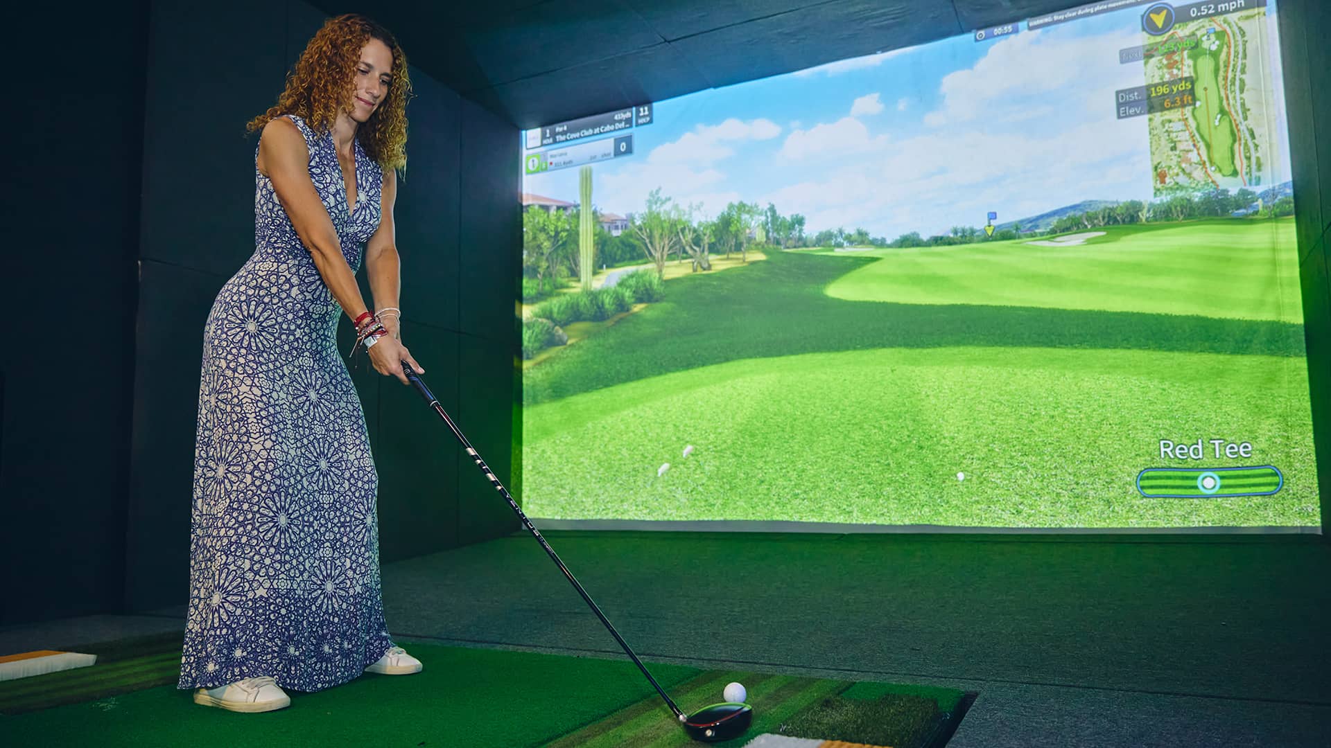 At a Mexico resort's conference event, a woman in a patterned dress holds a golf club, preparing to hit a ball on an indoor virtual golf simulator with a projected grassy course on the screen in front of her.