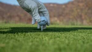 Primer plano de una mano enguantada colocando una pelota de golf en un tee en uno de los mejores complejos turísticos de México, con césped verde en primer plano y colinas marrones difuminadas al fondo; perfecto para eventos de conferencias o reuniones de hotel.