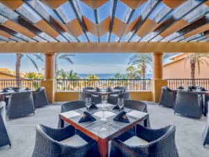 Terraza exterior de restaurante con sillas de mimbre y mesas bajo una pérgola de madera, perfecta para eventos o reuniones en complejos turísticos de México, con vistas a las palmeras y al océano a lo lejos en un día soleado.