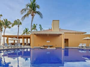 Una tranquila piscina al aire libre con agua cristalina de color azul, rodeada de tumbonas, sombrillas, palmeras y un edificio de estuco amarillo, perfecta para relajarse durante eventos de conferencias en los mejores complejos turísticos de México bajo un cielo azul y soleado.