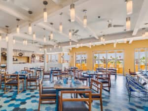 Un restaurante luminoso y moderno, perfecto para eventos y conferencias. Cuenta con mesas y sillas de madera, baldosas hexagonales azules en el suelo, paredes amarillas, grandes ventanales y numerosas lámparas colgantes en el techo. Las mesas están vestidas con servilletas y cristalería azules.