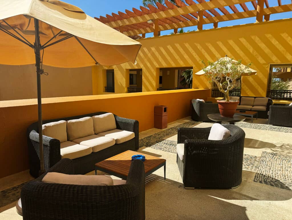 Outdoor patio at Mexico resorts features wicker furniture with beige cushions, a wooden table, and a large umbrella. Perfect for informal meetings or conference events, with potted plants and yellow walls casting striped shadows from the pergola overhead.