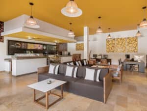 Interior de cafetería moderno con techo amarillo, lámparas colgantes, un sofá marrón con cojines blancos, mesas de madera y un mostrador con pasteles y carta de bebidas; ideal para reuniones informales o para relajarse después de eventos de conferencias en hoteles o complejos turísticos de México.