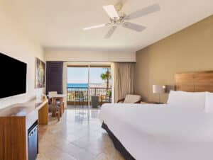 Una habitación de hotel moderna con una cama grande, muebles de madera, televisor de pantalla plana y una puerta corrediza de vidrio que da a un balcón con vista al océano; perfecta para relajarse después de eventos de conferencias en complejos turísticos de México.