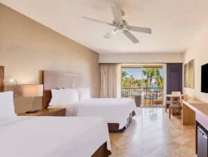 Una luminosa habitación de hotel con dos camas dobles, sábanas blancas, ventilador de techo, televisor, escritorio (ideal para reuniones) y balcón con vistas a palmeras y un paisaje soleado en uno de los mejores complejos turísticos de México.
