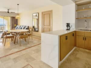 Espacio diáfano y moderno, ideal para reuniones, con zona de comedor con mesa y sillas de madera, sofá, espejo grande, acceso al balcón y cocina de madera clara con encimera de mármol. La luz natural inunda la estancia.