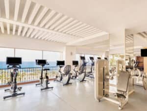 Un gimnasio luminoso y moderno en uno de los mejores complejos turísticos de México, con bicicletas estáticas y máquinas cardiovasculares frente a grandes ventanales con vistas a un soleado paisaje exterior, perfecto para los huéspedes que asisten a conferencias o reuniones.