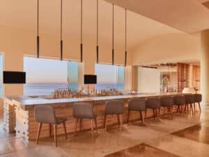 Un moderno bar con encimera de mármol, base de madera y sillas grises de respaldo alto, ideal para conferencias o reuniones en resorts mexicanos. Lámparas colgantes iluminan el techo mientras grandes ventanales ofrecen vistas al océano al atardecer. Detrás de la barra hay botellas y dos televisores de pantalla plana.