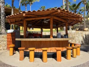 Un bar tiki redondo al aire libre, con techo de madera y taburetes, se ubica bajo las palmeras en uno de los resorts más exclusivos de México; ideal para relajarse después de conferencias o reuniones. Botellas y vasos relucen tras la barra bajo un cielo azul despejado.