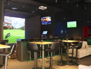 Un moderno salón de golf interior con mesas altas y taburetes de bar, grandes pantallas de simulador de golf que muestran campos de golf, varios televisores, un sofá e iluminación tenue: perfecto para reuniones informales o eventos de conferencias en los mejores hoteles y complejos turísticos de México.