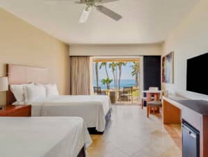 Una habitación de hotel en un complejo turístico de México con dos camas dobles, un televisor, un escritorio y una puerta corrediza de vidrio que da a un patio con asientos al aire libre, perfecta para relajarse después de eventos de conferencias, con vistas a palmeras y al océano.