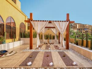 Zona de spa al aire libre con una pérgola de madera cubierta con cortinas blancas, esterillas de yoga y toallas en el suelo —perfecta para eventos de conferencias en complejos turísticos de México— rodeada de piedras blancas, cactus en macetas y edificios amarillos bajo un cielo azul despejado.
