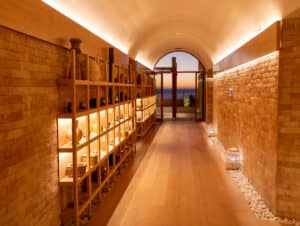 Un pasillo cálido y luminoso con suelos de madera y paredes de ladrillo, con estanterías decorativas a la izquierda. Al final, unas puertas de cristal dan a una vista del océano y de una vibrante puesta de sol, ideal para reuniones memorables en los resorts mexicanos. Faroles y piedras blancas adornan los bordes del suelo.