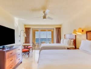 Una luminosa habitación de hotel en uno de los mejores resorts de México, con dos camas dobles, un gran televisor de pantalla plana, una mesita con sillas y un balcón con vista al mar: ideal para relajarse después de eventos o reuniones. La iluminación cálida contribuye a crear un ambiente acogedor.