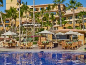 Mesas y sillas de madera con cristales verdes están dispuestas bajo sombrillas junto a una piscina de azulejos azules en uno de los mejores complejos turísticos para conferencias y eventos de México, con palmeras y un edificio de hotel de color tostado al fondo, bajo un cielo azul despejado.