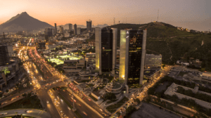 Vista aérea de una ciudad al atardecer con edificios altos y modernos, calles concurridas iluminadas por las luces de los coches y una gran montaña al fondo: un escenario ideal para conferencias y reuniones en los mejores hoteles de México.