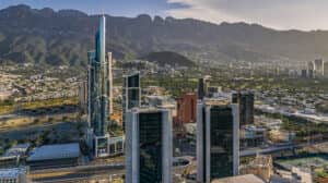 Vista aérea de una ciudad moderna con altos rascacielos y una distintiva torre de cristal, que alberga los mejores hoteles de México, perfectos para conferencias y reuniones, con verdes montañas de fondo bajo un cielo despejado.