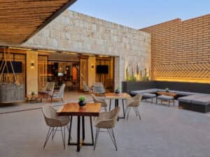 Patio exterior con mesas de madera, sillas de mimbre, plantas en macetas y bancos acolchados, ideal para reuniones informales o eventos de conferencias. Muros de piedra y ladrillo enmarcan una vista despejada hacia un moderno salón interior en uno de los resorts más exclusivos de México.