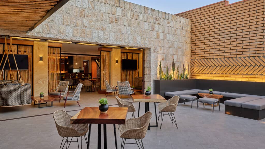 Modern outdoor patio with wooden tables, wicker chairs, gray cushioned benches, potted plants, and a stone wall—ideal for conference events or meetings. The area opens to an indoor bar with warm lighting and natural wood accents.