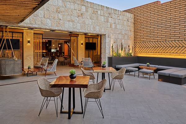 Modern outdoor patio at Mexico hotels with wooden tables, wicker chairs, grey cushioned benches, potted plants, and warm lighting against stone and brick walls—perfect for meetings or conference events, opening into a stylish indoor lounge area.