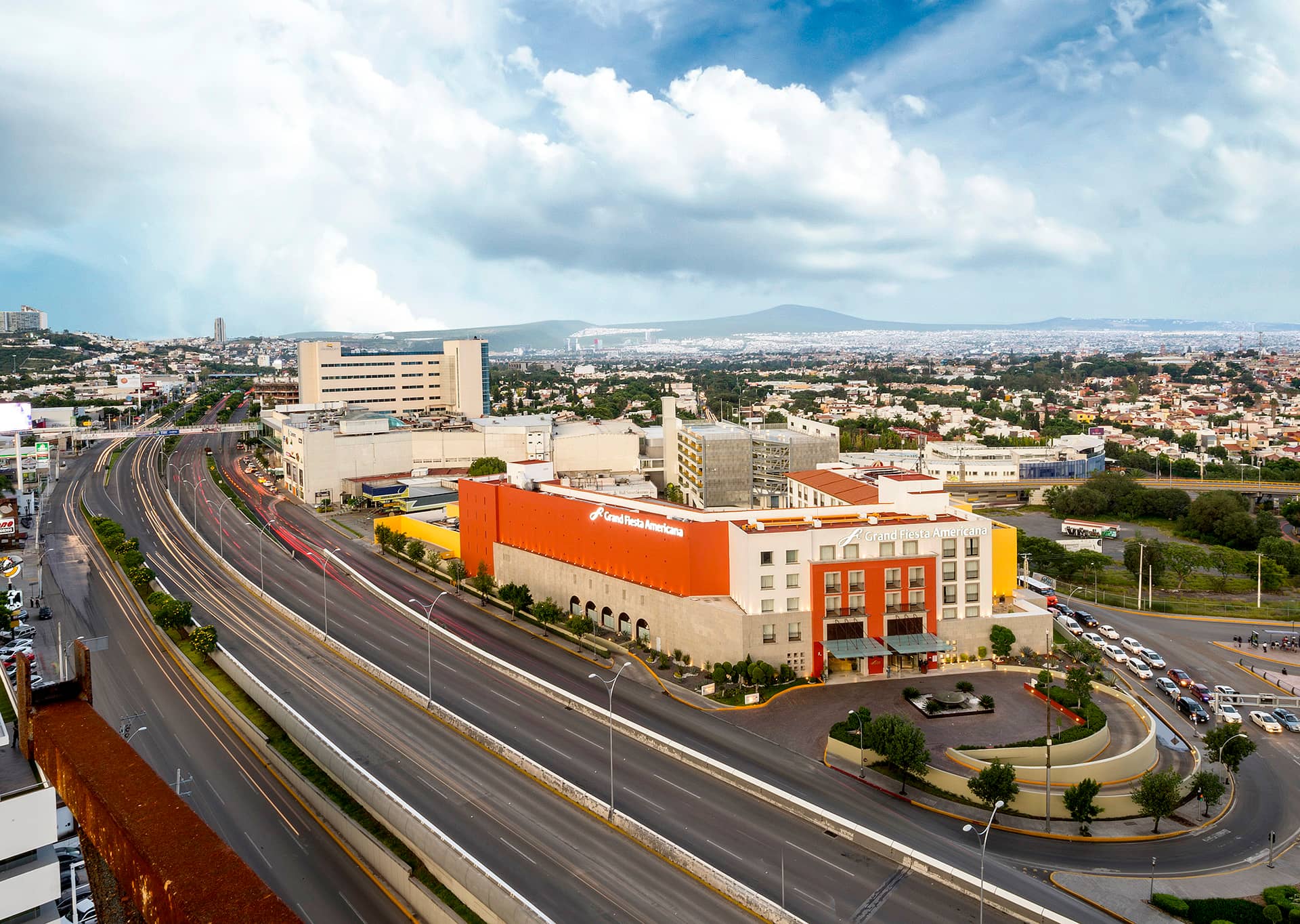 Aerial view of a large city in Mexico with a prominent orange and white hotel near wide, curving roads, surrounded by many smaller buildings—perfect for meetings and conference events under a cloudy blue sky.