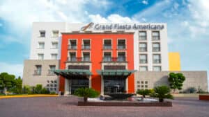 Vista frontal del hotel Grand Fiesta Americana en México, que destaca por su arquitectura moderna con paredes rojas, blancas y beige, grandes ventanales, jardines paisajísticos y una fuente redonda: un entorno ideal para reuniones y conferencias bajo un cielo azul brillante.