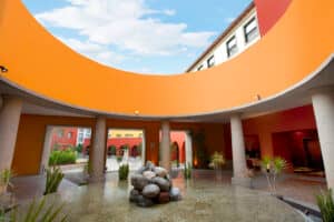 Un patio circular en un resort mexicano cuenta con un techo curvo de color naranja, columnas de piedra y una fuente central con rocas, rodeada de pequeñas plantas. El cielo azul y las coloridas paredes del hotel crean un entorno perfecto para reuniones o conferencias.