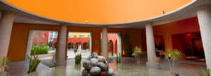 Patio circular con altas columnas, techo curvo de color naranja, fuente central de piedra sobre una superficie de agua plana y plantas en macetas a su alrededor: un entorno acogedor para eventos y conferencias en hoteles y resorts de lujo en México. Se aprecian edificios y arcos de colores vivos.