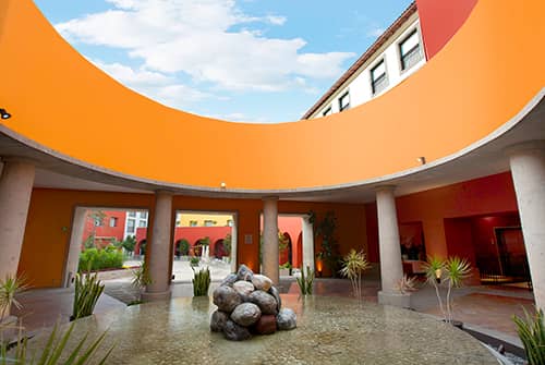 A circular courtyard with orange walls and columns surrounds a shallow pool, perfect for meetings or conference events at resorts in Mexico. Green plants line the water’s edge beneath a blue sky with scattered clouds.