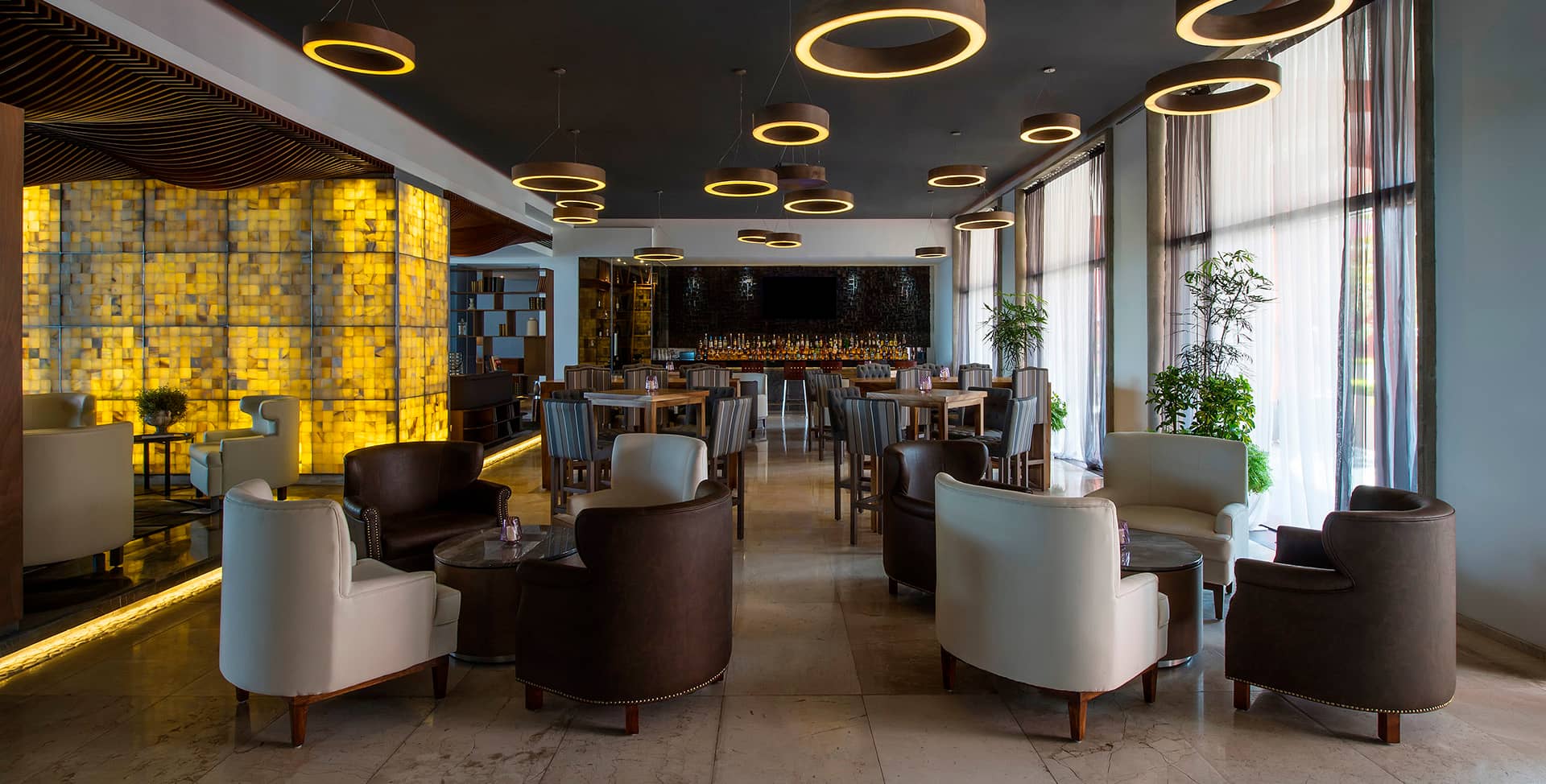 Modern lounge in Mexico resorts features cream and brown armchairs around small tables, large windows with sheer curtains, circular ceiling lights, and a backlit wall—an inviting space perfect for meetings or conference events, with plants adding a touch of greenery.