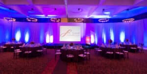 Un salón de baile acondicionado para eventos de conferencias con mesas redondas, elegantes arreglos de mesa, centros de mesa florales y un escenario con una gran pantalla que muestra el logotipo de Grand Fiesta Americana, iluminado con luces azules y moradas.