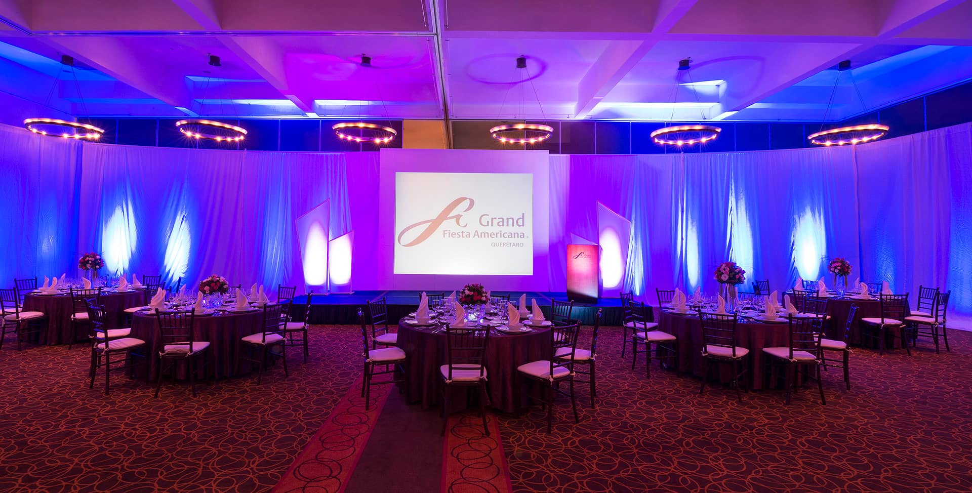 A ballroom set for conference events with round tables, elegant place settings, floral centerpieces, and a stage with a large screen displaying the Grand Fiesta Americana logo, illuminated with blue and purple lights.