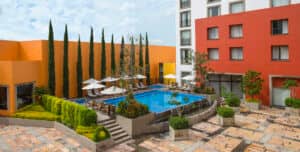 Una pequeña piscina exterior de hotel rodeada de tumbonas y sombrillas, bordeada por árboles altos y esbeltos y muros de color naranja, con un moderno edificio de varias plantas al fondo: perfecta para reuniones o eventos de conferencias en los mejores complejos turísticos de México.