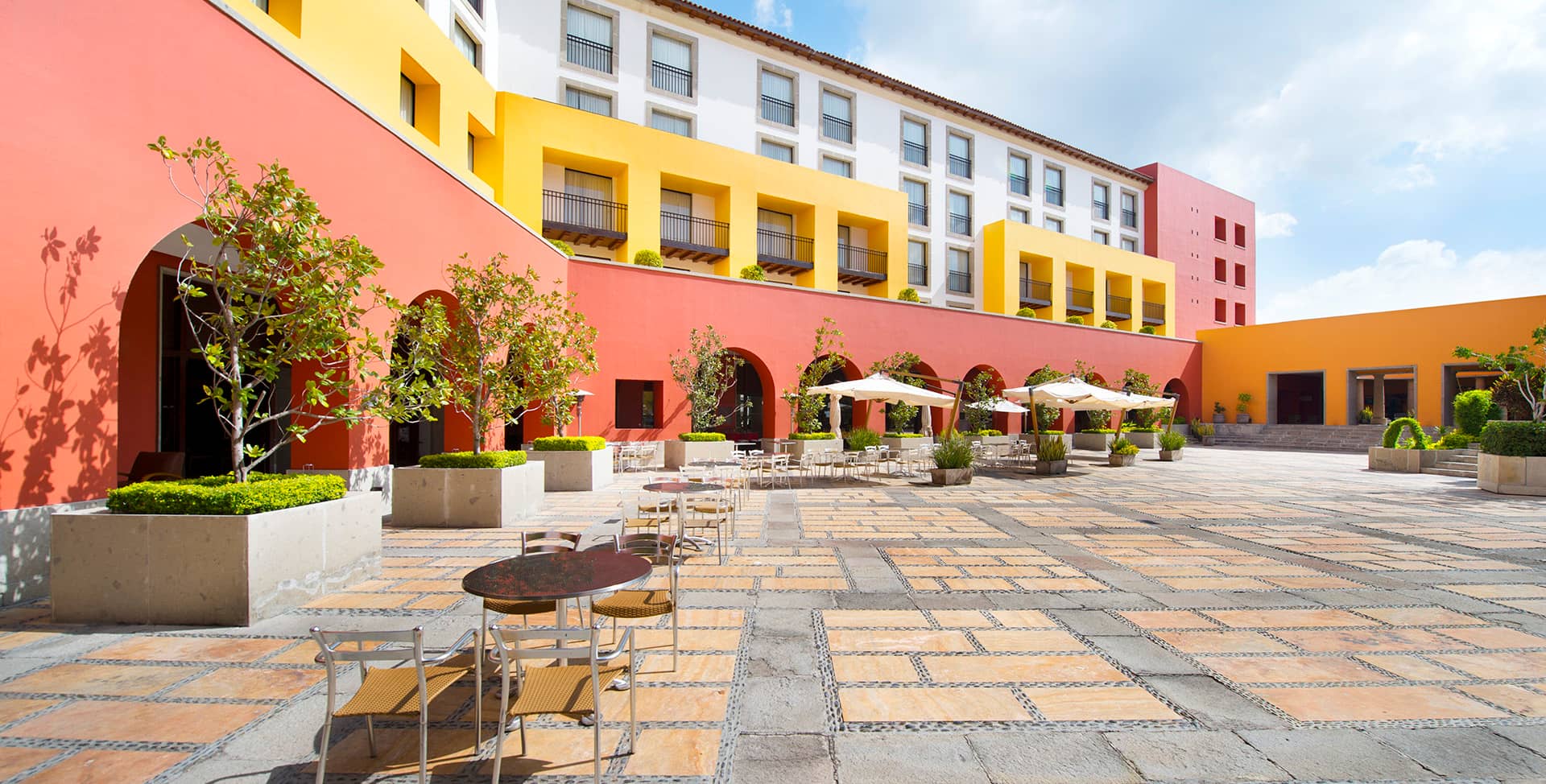 A spacious outdoor courtyard with patterned stone tiles, red and yellow building walls, potted plants, and seating areas—perfect for conference events or relaxed meetings at resorts and hotels in Mexico under a bright blue sky.