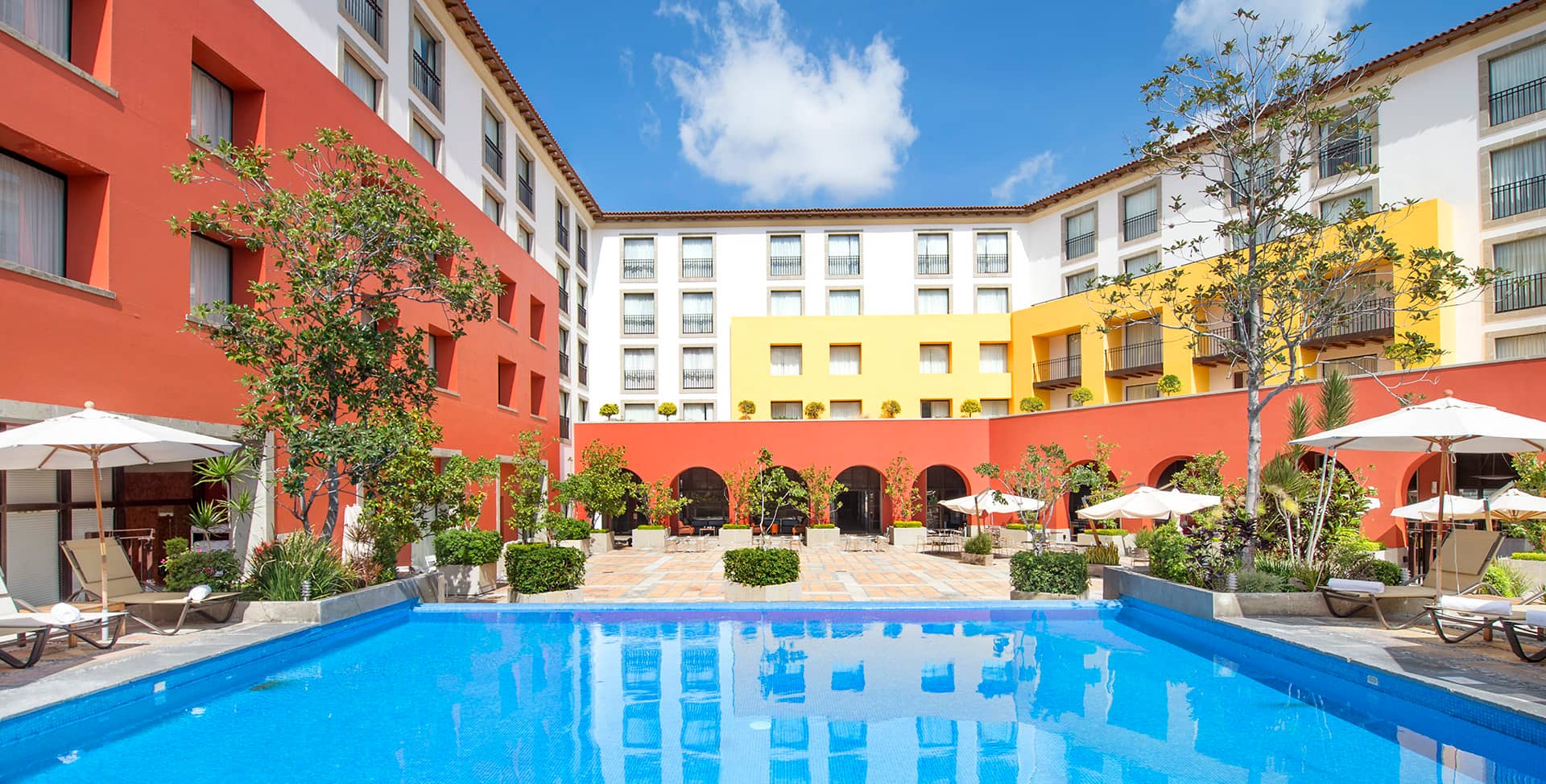 A vibrant hotel courtyard at one of Mexico's finest resorts, featuring red and yellow walls, arched walkways, poolside lounge chairs, lush green plants, and a clear blue swimming pool—perfect for meetings or conference events.