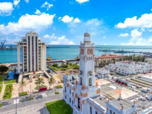 Vista aérea del puerto de Veracruz, México, que muestra edificios blancos históricos, palmeras y una torre del reloj junto al muelle: un entorno acogedor para eventos o reuniones cerca de reconocidos complejos turísticos y hoteles, con el océano y el cielo azul de fondo.