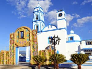 Una iglesia blanca con detalles azules se alza bajo un cielo radiante, ideal para conferencias o reuniones. Frente a ella, una entrada arqueada está decorada con coloridas flores, mientras que palmeras y una farola de estilo retro realzan el ambiente del complejo.