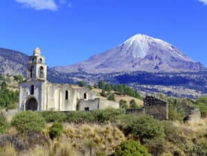 En primer plano se alza una antigua iglesia abandonada, cubierta de vegetación, mientras que al fondo se eleva majestuosamente una montaña volcánica nevada: una vista inspiradora para eventos de conferencias o reuniones inolvidables en los mejores complejos turísticos de México.