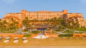 Un gran complejo turístico naranja frente al mar en México, con balcones, palmeras, piscinas y cabañas con techo de paja —perfecto para reuniones o eventos de conferencias—, se encuentra frente a una playa de arena bordeada de tumbonas y sombrillas bajo un cielo azul despejado.