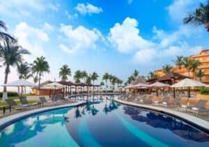 Una lujosa piscina estilo resort con tumbonas y sombrillas, rodeada de palmeras y edificios del complejo bajo un cielo azul brillante: perfecta para celebrar reuniones o eventos memorables en los mejores hoteles de México.